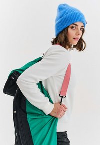 Mladá žena v modrej pletenej čiapke a bielej svetri nesie zeleno-čiernu bundu s ružovým popruhom cez rameno.