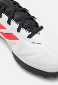 Botas de fútbol blancas con acentos negros, parte superior sintética texturizada, detalles rosados y suela de goma negra con tacos. La etiqueta dice "FUSIONFEEL."