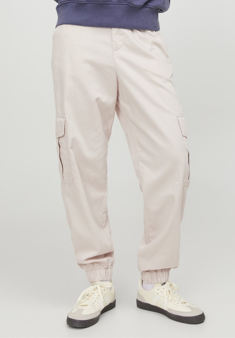 Světle růžové cargo kalhoty s elastickými manžetami a dvěma bočními kapsami. Vyrobeny z hladké, odolné tkaniny. Nosič má na sobě bílé tenisky.