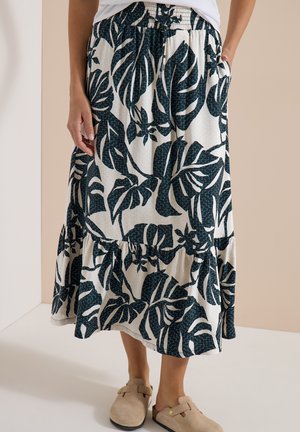 Femme portant une jupe blanche avec un grand imprimé de feuilles tropicales vert foncé, taille élastique et chaussures beiges sans lacets.