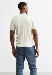 Chandail tricoté à manches courtes en crème, featuring des côtes verticales et un ourlet ajusté. Associé à un pantalon rayé bleu, montrant la vue arrière.