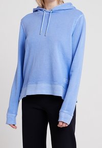 Sudadera con capucha de color azul claro hecha de una tela suave, con mangas largas, un ajuste relajado y una etiqueta con logo en la parte delantera. Costuras limpias y cordones.