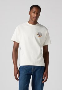 Balta medvilninė marškinėliai su apvaliu kaklu, trumpomis rankovėmis ir spalvinga "LEVI'S" grafika ant krūtinės. Dėvimi su mėlynais džinsais.