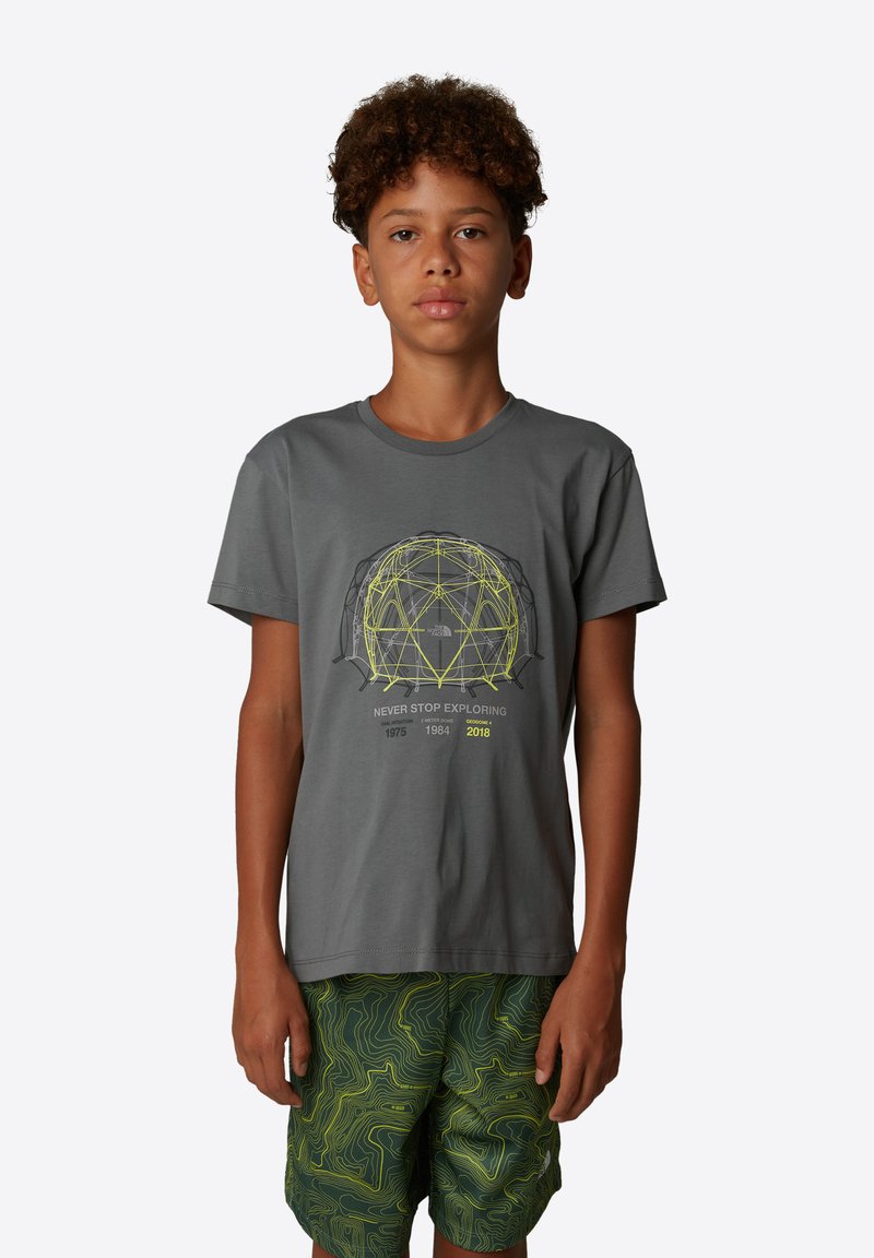 Camiseta de algodón gris con cuello redondo y mangas cortas, con un gráfico geométrico amarillo y texto. Combinada con pantalones cortos verdes de patrón ondulado.
