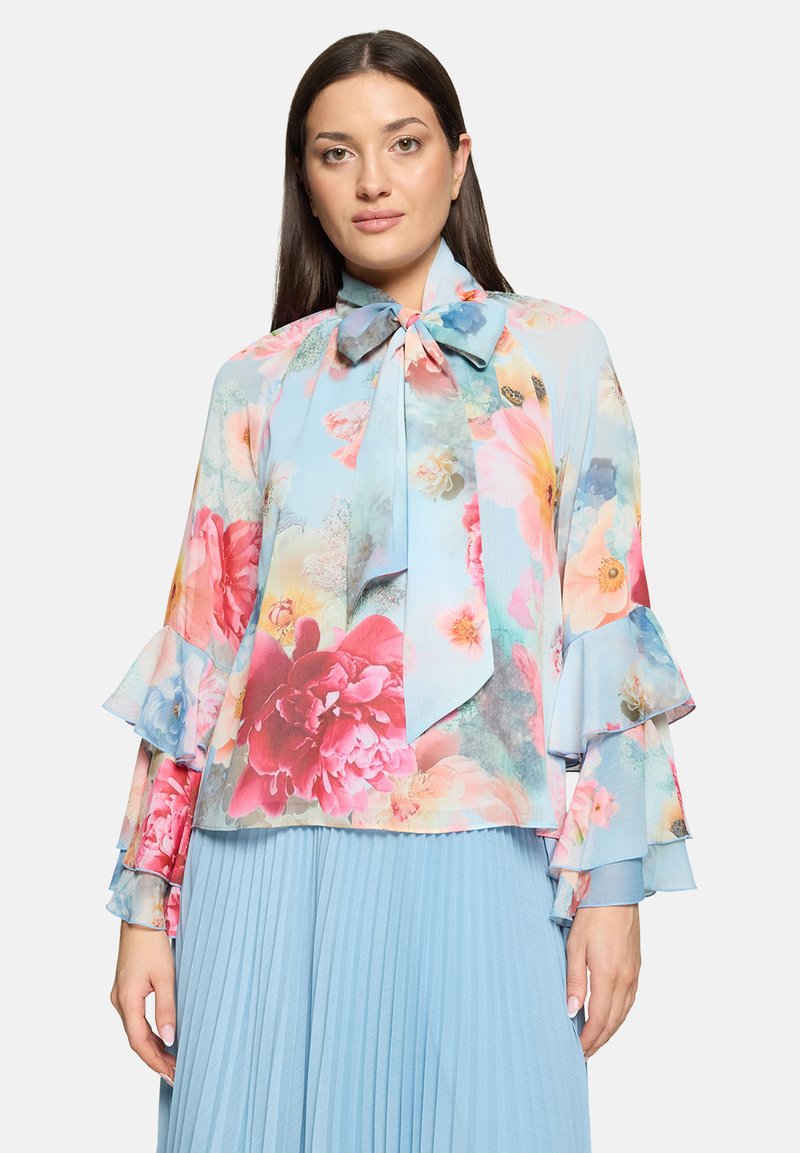Blouse fleurie bleu clair, avec un col à nœud papillon et des manches longues à volants. Confectionnée dans un tissu transparent orné d'un motif floral éclatant.