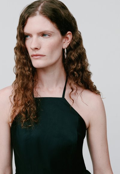 Femme aux longs cheveux bruns bouclés, portant un haut noir à col halter et des boucles d’oreilles en argent en forme d’anneau, regardant sur le côté sur un fond gris uni.