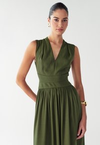 Zelena haljina bez rukava s V-izrezom, prilagođenim korzetom i lepršavom suknjom. Ima suptilnu teksturu i nabore na struku, stvarajući lepršavu siluetu.