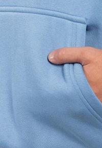 Sudadera de tejido suave en azul claro, con un bolsillo frontal. Costura visible a lo largo del bolsillo y el escote. Mano descansando en el bolsillo.