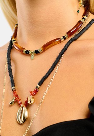 Trois colliers superposés : un avec des perles couleur ambre, un autre avec des disques en caoutchouc noir, et un troisième avec des coquillages dorés et des pierres précieuses rouges.