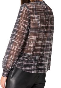 Femme portant un chemisier transparent à manches longues avec un motif abstrait en grille rose, noir et blanc, associé à un pantalon en cuir noir.