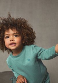 Türkiissinine puuvillane dressipluus soonikkoes mansettide ja kaelusega; rinnal väike tikitud muster. Käharad juuksed ja hele nahatoon.