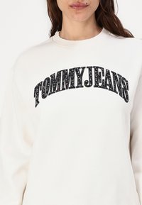 Kremfarget genser med en svart, teksturert "TOMMYJEANS" logo over brystet. Rund hals og lange ermer med en avslappet passform.