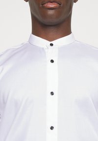 Uomo che indossa una camicia bianca con colletto alla mandarino e bottoni neri, mostrato dalle spalle alla bocca su uno sfondo bianco.