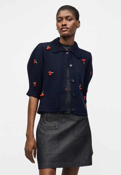 Γυναίκα που φοράει ένα σκούρο μπλε πουλόβερ με κόκκινη κεντητή κερασιά και μια σκούρα γκρι φούστα, στέκεται μπροστά σε απλό ανοιχτό φόντο.