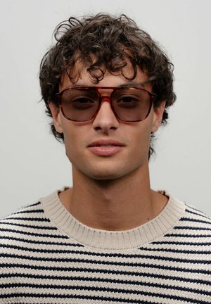 Lunettes de soleil oversize marron avec une monture transparente, portées par une personne dans un pull crème et marine à rayures avec un col côtelé.