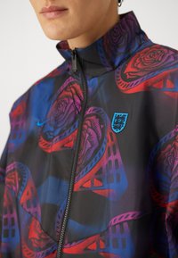 Windbreaker jacket in a black fabric featuring a multicolor rose pattern. High collar, zip front, and a blue logo patch on the chest.