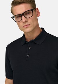 Homme portant des lunettes écaille de tortue noires et un polo noir à manches courtes avec deux boutons attachés, regardant vers la caméra.