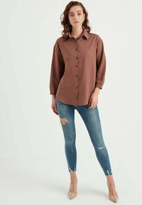 Camicia marrone con bottoni e colletto classico, abbinata a jeans blu strappati e décolleté beige. Tessuto morbido e vestibilità rilassata.