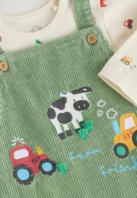 Salopette en velours côtelé vert avec des motifs de vache et de tracteur brodés. Boutons en bois et chemise douce couleur crème avec des motifs de véhicules colorés.
