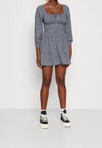 Námořnická modrá květinová šaty s nabíranými rukávy a čtvercovým výstřihem; nošeno s černými vysokými teniskami s bílými šňůrkami a gumovou podrážkou.