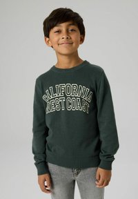 Zeleni džemper s bijelim natpisom "CALIFORNIA WEST COAST", okruglim izrezom, rebrastim manžetnama i rubom; upareno sa sivim trapericama.