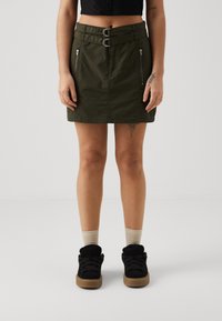 Grønn skjørt med to sølvspenne detaljer i midjen, to glidelåslommer, og rett snitt. Iført med svarte sko og beige sokker.