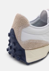 Gros plan du talon d'une basket en daim beige, cuir blanc et semelle en caoutchouc bleu marine avec des crampons circulaires en relief pour l'adhérence.