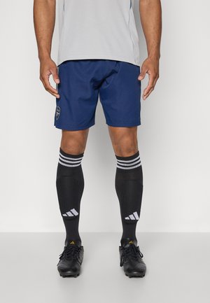 Marine sports shorts med logo, parret med sorte sokke-lignende benklæder med hvide striber og branding, samt sorte fodboldstøvler.