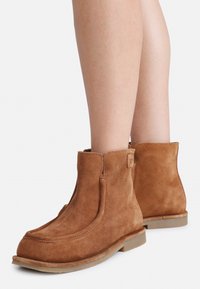 Braune Wildleder-Stiefeletten mit abgerundetem Zehenbereich, seitlichen Nähten und niedrigen Gummisohlen, die eine weiche Textur und ein minimalistisches Design aufweisen.