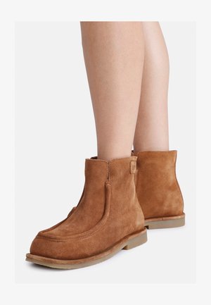 Botas de tornozelo em camurça castanha com bico arredondado, costuras laterais e solas de borracha baixas, apresentando uma textura suave e um design minimalista.