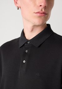 Juodas polo marškinėliai, pagaminti iš medvilnės; turi klasikinį apykaklę, trijų mygtukų užsegimą ir subtilų iškilmingą logotipą kairėje krūtinės pusėje.