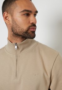Sweatshirt zippé beige avec un col haut, texture côtelée et un petit logo brodé sur la poitrine. Finition en tissu lisse.