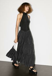 Top nero con collo all'americana abbinato a una gonna maxi nera a strati e fluida. La modella tiene una pochette bianca e indossa sandali neri con zeppa.