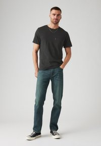 Vyras stovi su viena ranka kišenėje, vilkėdamas juodą marškinėlį, mėlynas džinsus, juodus sportbačius ir ploną auksinę grandinėlę ant paprasto fono.