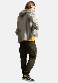 Giacca a zip grigio chiaro con cappuccio e polsini elastici, abbinata a pantaloni cargo verde oliva con molteplici tasche e sneakers nere.