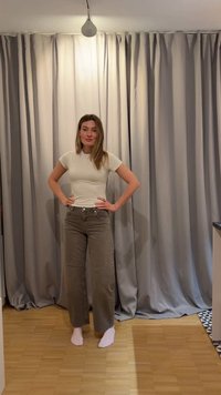 Femme debout pieds nus sur un sol en bois, portant un t-shirt blanc et un pantalon marron à jambes larges devant des rideaux gris fermés.