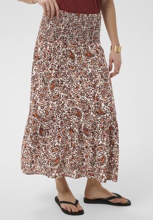 Flydende ankel-lang nederdel med brun og orange blomstret print, smock-talje, båret med sorte klipklappere og guldsmykker på en hvid baggrund.