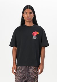 Person med langt svart hår iført en svart Adidas t-skjorte med basketballgrafikk og leopardmønstrede bukser mot en hvit bakgrunn.