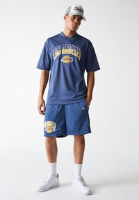 Mėlynas Sportinis marškinėlis su tinklu, ant kurio užrašas LOS ANGELES LAKERS, derinamas su atitinkančiomis šortais. Baltos sportinės bateliai ir kepurė puikiai užbaigia aprangą.