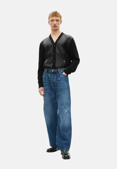 Ung mand iført en sort knap cardigan med læderpaneler, løse blå jeans og sorte sko, som står med den ene hånd i lommen.