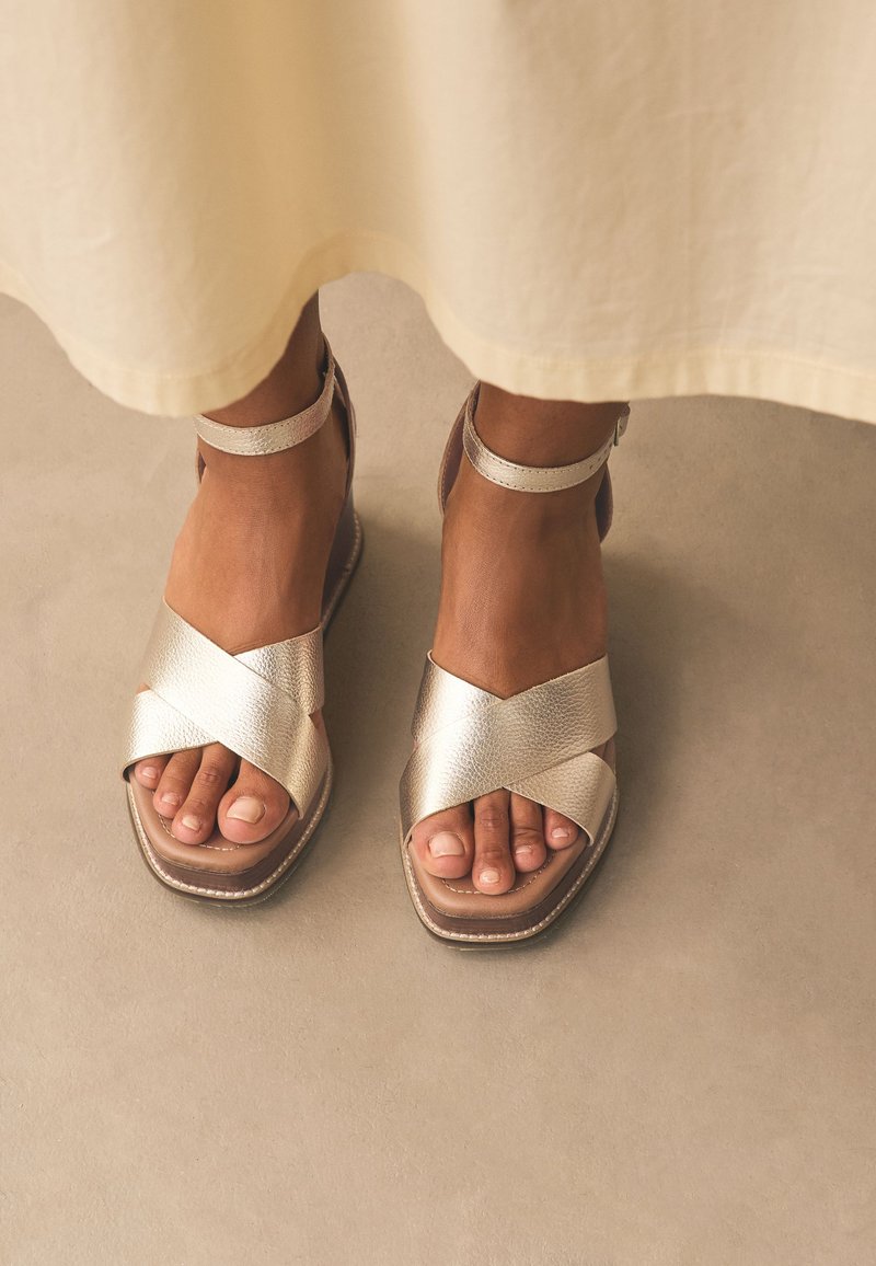 Fémes arany színű pántos szandálok széles keresztpánt dizájnnal, lapos talppal és bokapánttal; texturált anyag sima felületekkel.