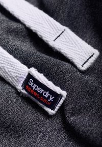 Tela gris con una superficie texturizada; presenta una etiqueta blanca con el logo "Superdry" en negro y rojo, con detalles de costura.