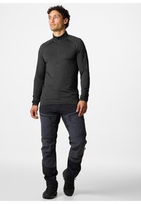 Maglietta nera a maniche lunghe con colletto a mezza zip; abbinata a pantaloni grigio scuro con numerose tasche con zip e robusti stivali neri.