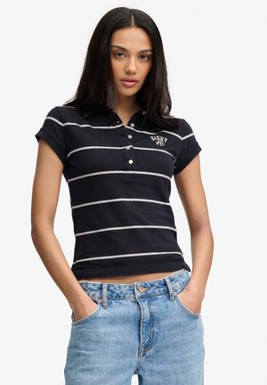 Joven mujer de pie con las manos en los bolsillos, vistiendo una camiseta polo negra a rayas y vaqueros azul claro sobre un fondo liso.