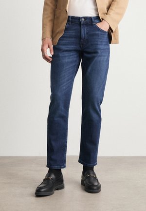 Jeans in denim blu scuro con taglio dritto, dotati di tasche anteriori, chiusura con bottone e cuciture minimali. Abbinati a mocassini neri.