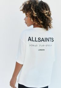 Bílé oversized tričko s krátkými rukávy. Na zádech se nachází černý text s logem: "ALLSAINTS" a "LONDÝN", doplněný textem v japonštině.