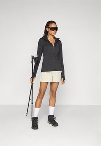 Zwarte zip-up jas, lichtbeige shorts, zwarte wandelschoenen en witte sokken. Houdt trekkingstokken vast. Glad materiaal, sportief ontwerp, outdoor kleding.