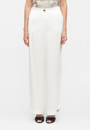 Nő magas derekú, bő szárú fehér nadrágot visel oldalzsebekkel és fekete szövött nyitott orrú cipőkkel, egyszínű háttér előtt áll.