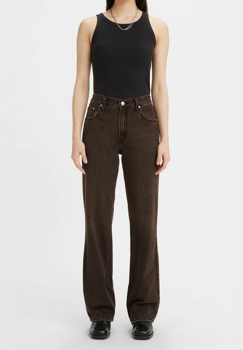 Femme portant un haut noir sans manches, un jean droit taille haute marron foncé, des chaussures noires et un collier chaîne argenté, debout devant un fond blanc.