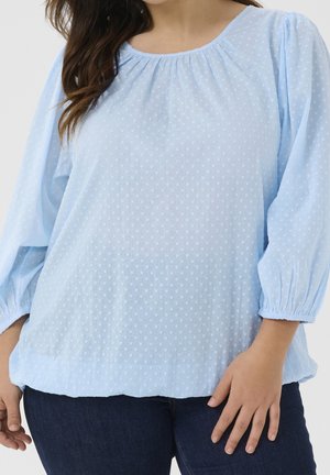 Woman wearing light blue textured blouse with gathered neckline and three-quarter sleeves, paired with dark blue jeans.